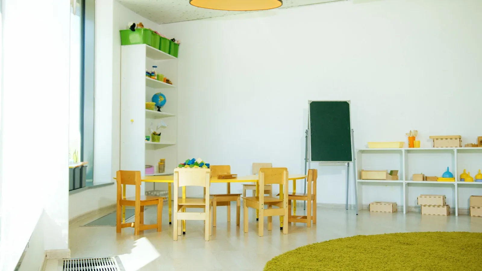 An empty, brightly lit kindergarten classroom with small wooden chairs around a low table, a green chalkboard on an easel, and shelves of toys and learning materials.