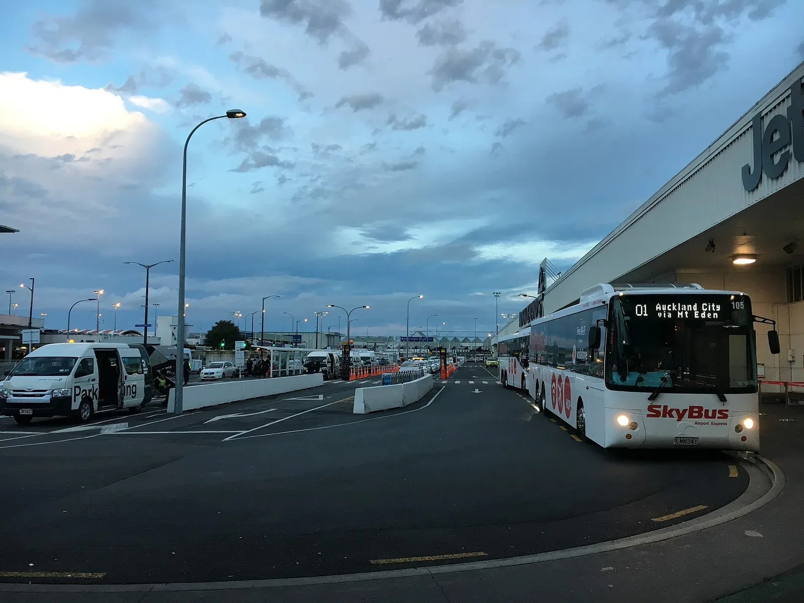 Kerbside outside a low airport terminal building at dusk, with a white SkyBus coach and minibuses parked along the road under street lights
