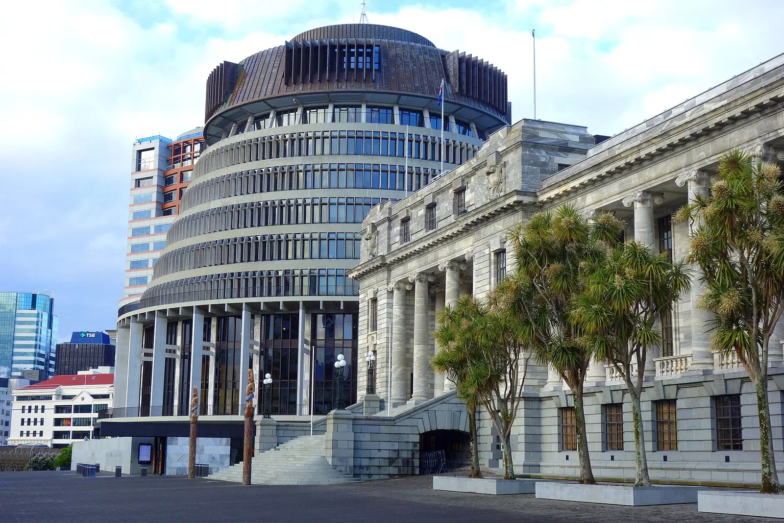 The circular Beehive building beside the columned stone facade of Parliament House, with cabbage trees in front, under a cloudy sky