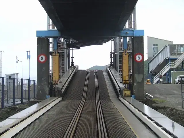 Looking down a steel loading ramp with three interlaced rail tracks set into it, passing beneath a blue overhead gantry structure, with a raised road-deck walkway and terminal buildings to the side