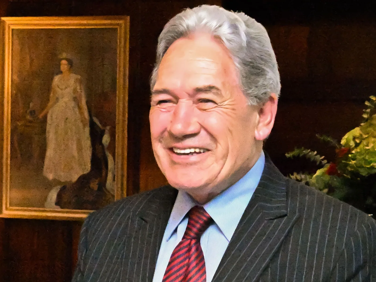 Winston Peters, smiling, in a pinstriped suit and red tie, in a wood-panelled room with a framed portrait behind him