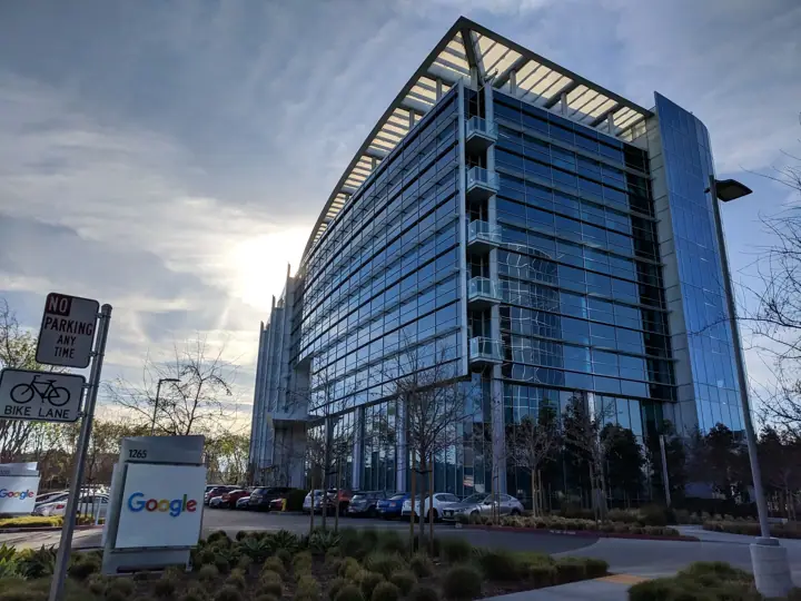 A tall glass-walled office building under a cloudy sky, with a Google sign on a low monument in the landscaped area in front