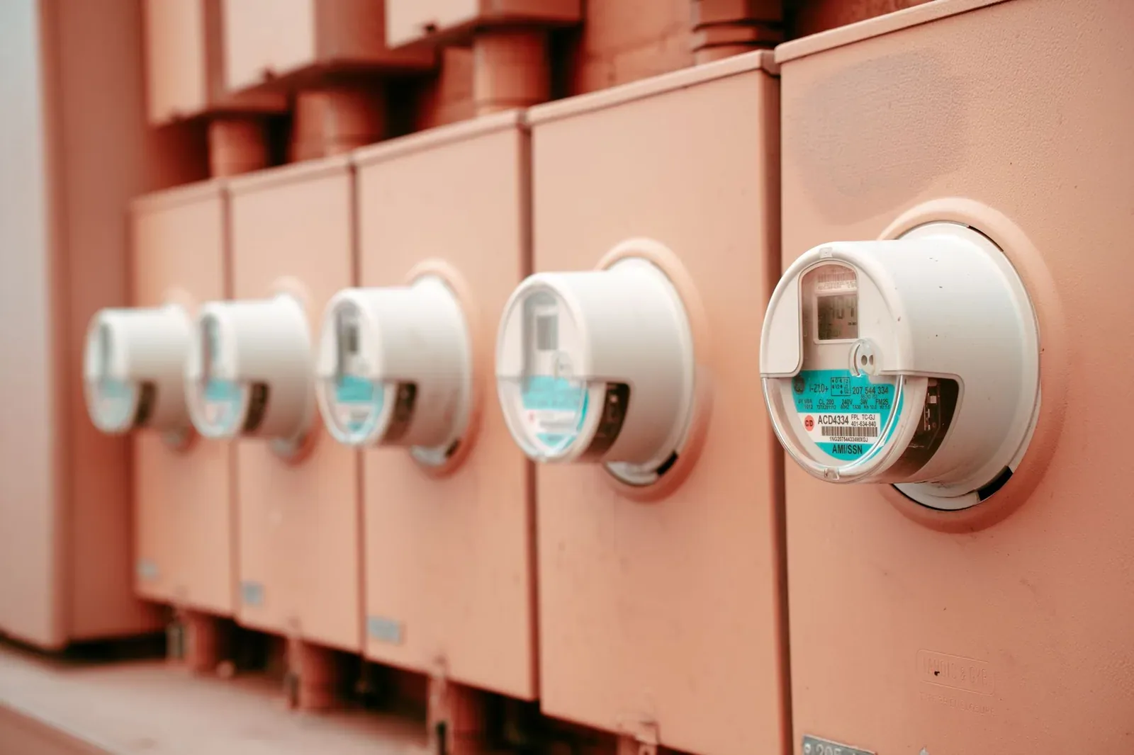 A row of white electricity meters mounted on pinkish metal cabinets, seen at an angle with the nearest meter in focus