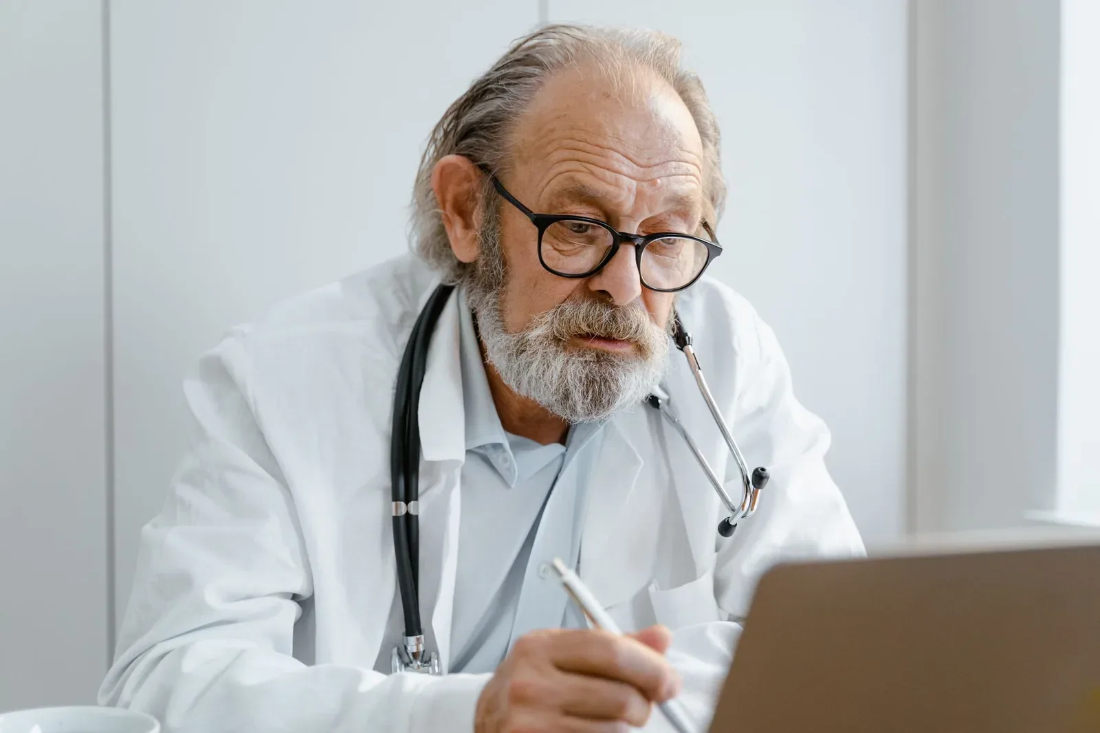 An older doctor with grey hair, a beard, glasses and a stethoscope, wearing a white coat, writing notes while looking at a laptop screen