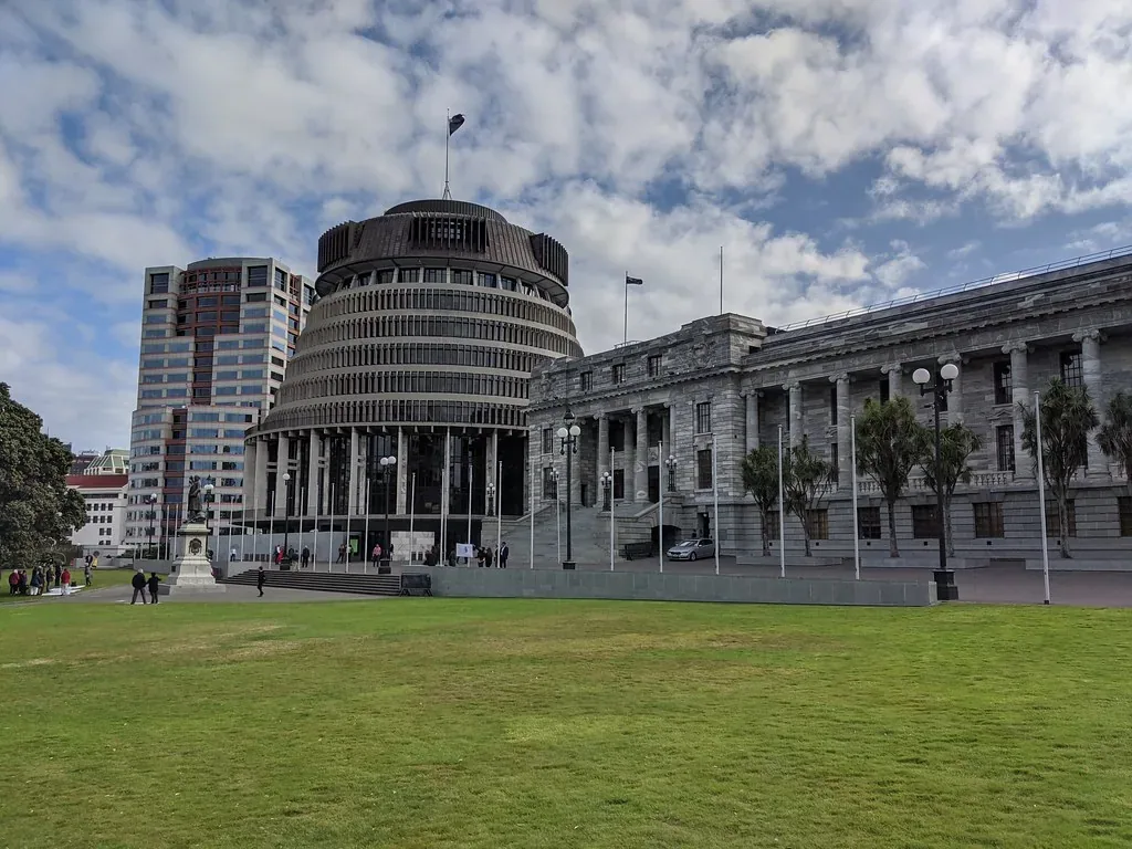 The round, tiered Beehive building beside the stone Parliament House, with a lawn in front and a New Zealand flag flying on the roof