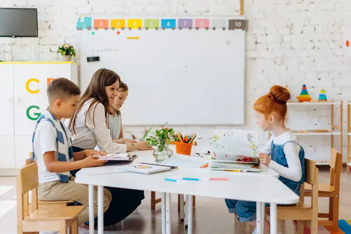 A teacher sits at a white table with three young pupils looking at open books, while another pupil reads at the end of the table in a bright classroom with a whiteboard and coloured number cards on the wall.