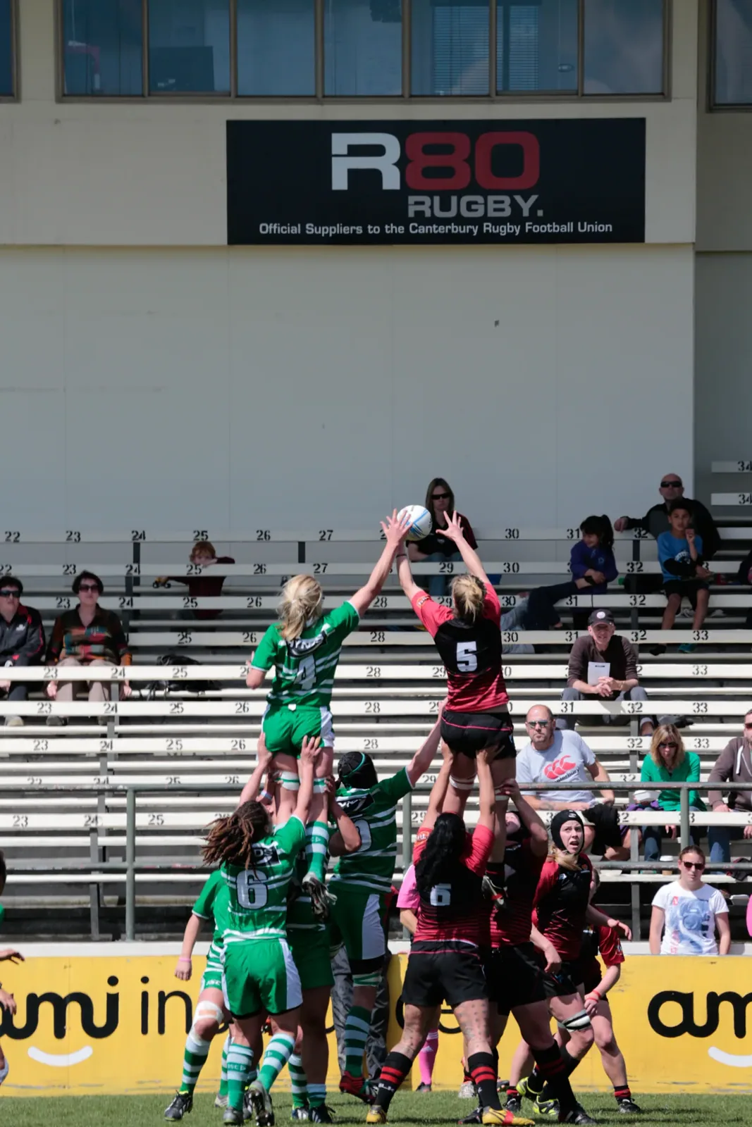 Women's rugby players contest a lineout, one player in green and one in red and black lifted high reaching for the ball, with spectators in the stand behind