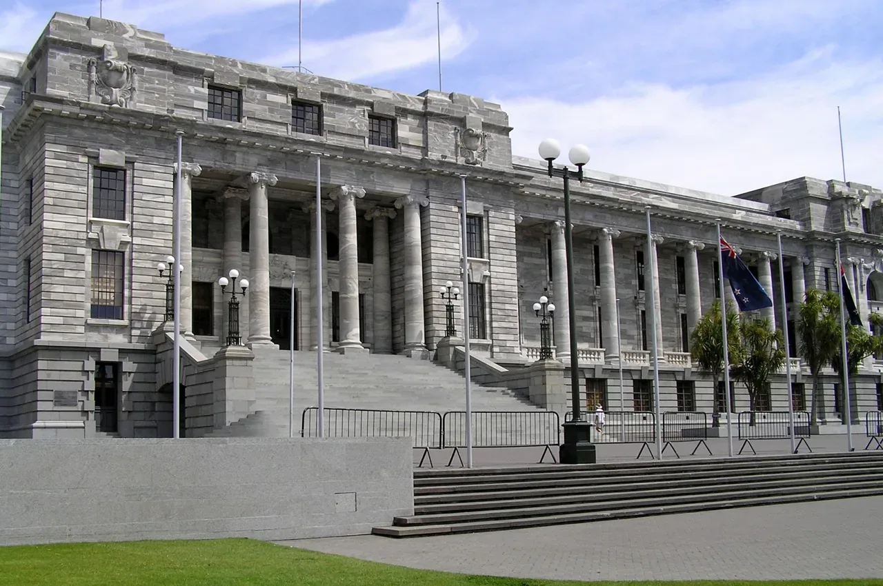 The grey stone colonnaded facade and front steps of Parliament House in Wellington under a blue sky, with the New Zealand flag flying at right