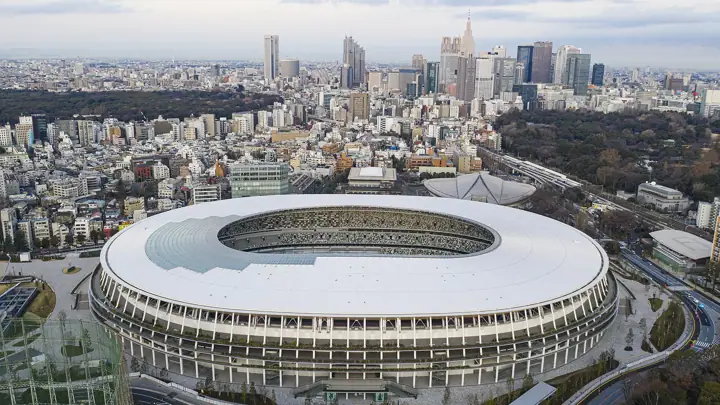 Aerial view of a large oval stadium with a white roof ring and open centre, surrounded by dense city blocks and high-rise towers