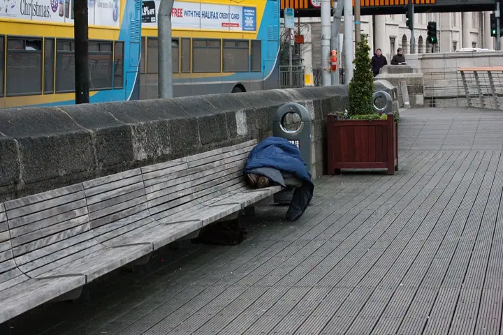 A person curled up under a blue blanket, sleeping on a wooden bench beside a stone wall on a city walkway, with a double-decker bus passing behind