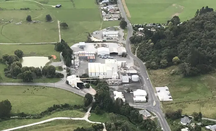 Aerial view of a large dairy processing plant with white buildings and storage tanks, surrounded by green farmland, trees and a winding road
