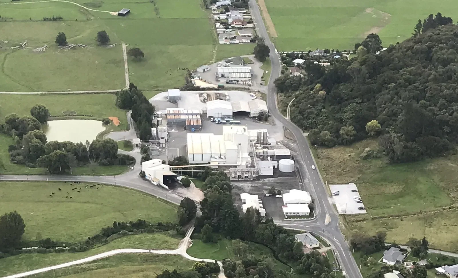 Aerial view of a large dairy processing plant with white buildings and storage tanks, surrounded by green farmland, trees and a winding road