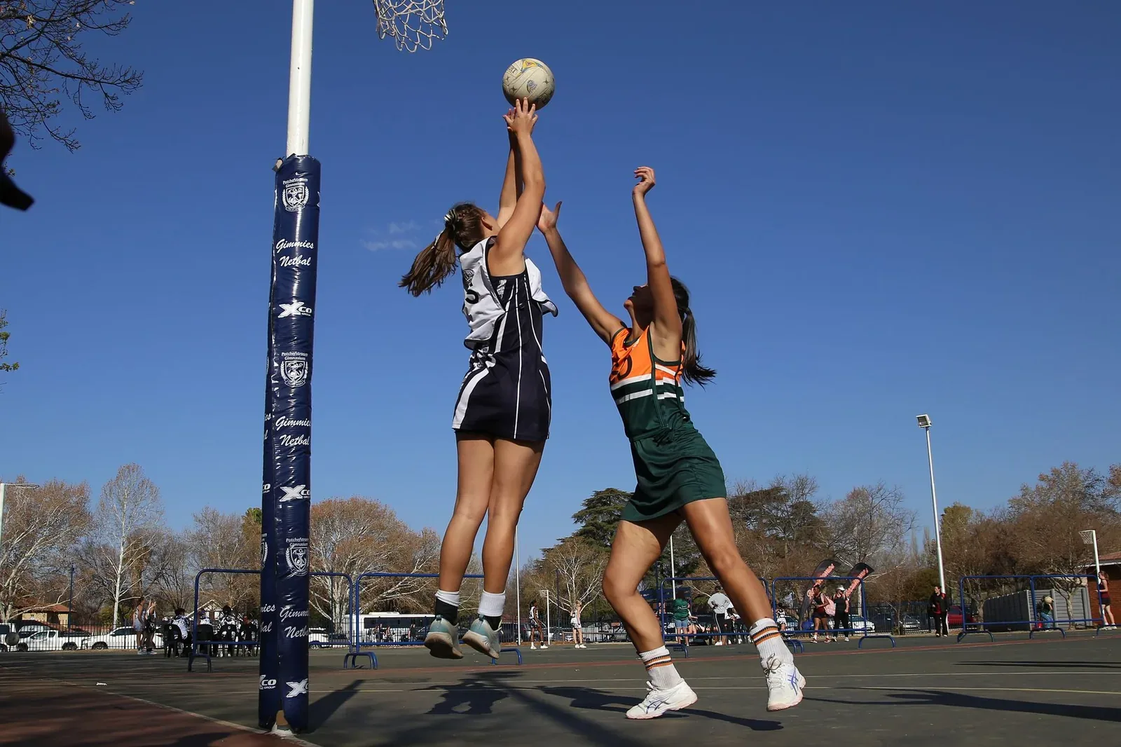 Two netball players leap for the ball beside a netball goal post on an outdoor court under a clear blue sky