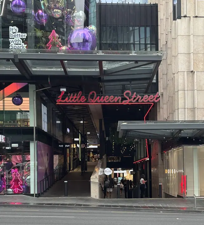 The street entrance to Little Queen Street at Commercial Bay Shopping Centre in Auckland, with a red neon sign over a covered walkway lined with shops and Christmas decorations in the windows above