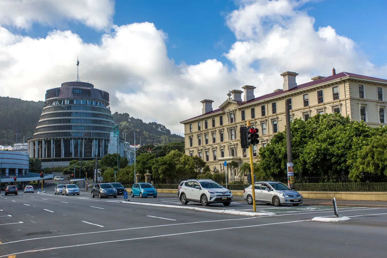 The circular Beehive building and a historic cream-coloured government building beside a road in Wellington under a partly cloudy sky