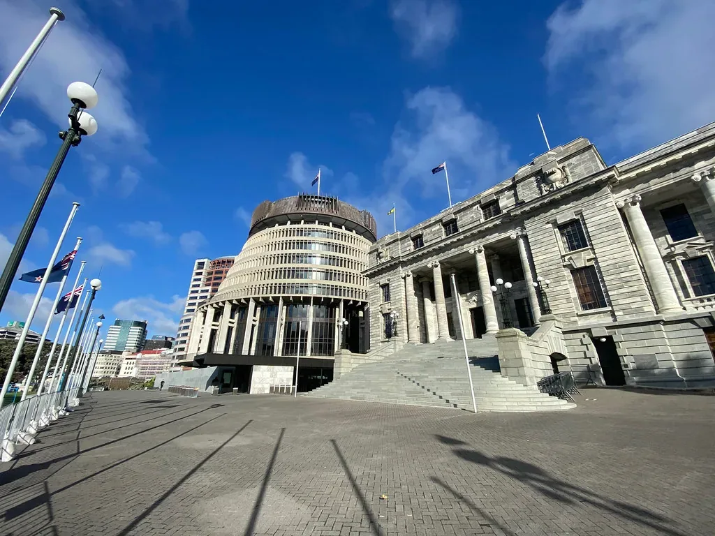 Low-angle view across a forecourt with flagpoles of the circular Beehive building beside the columned stone Parliament House under a blue sky