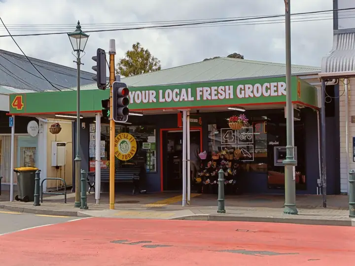 A single-storey corner grocery shop with a green fascia reading 'Your local fresh grocer', flowers displayed outside, beside a traffic light on a quiet street.