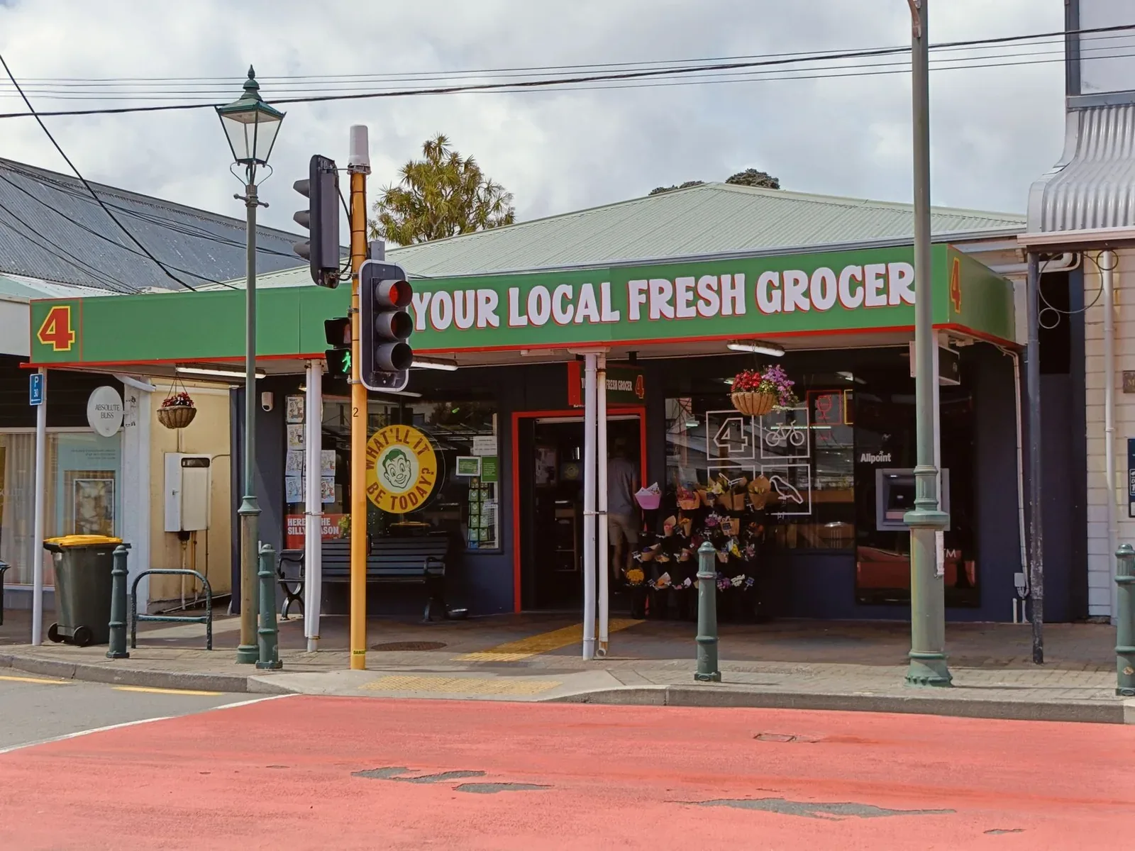 A single-storey corner grocery shop with a green fascia reading 'Your local fresh grocer', flowers displayed outside, beside a traffic light on a quiet street.