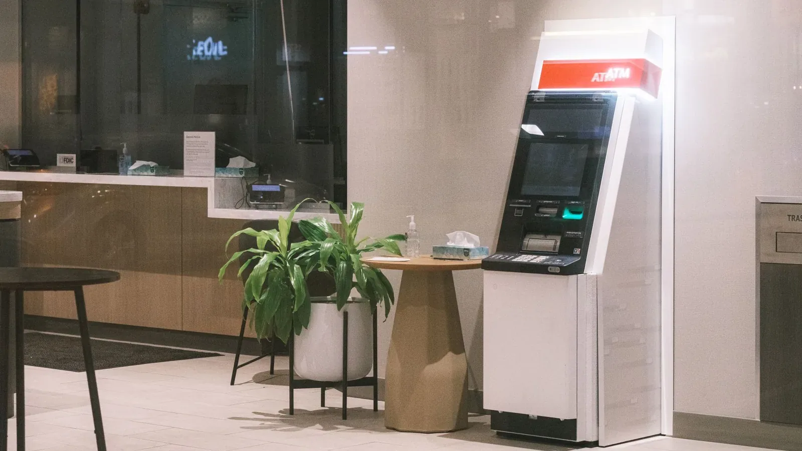 A freestanding ATM with a red 'ATM' sign next to a potted plant and a small round table in a modern, quiet bank lobby