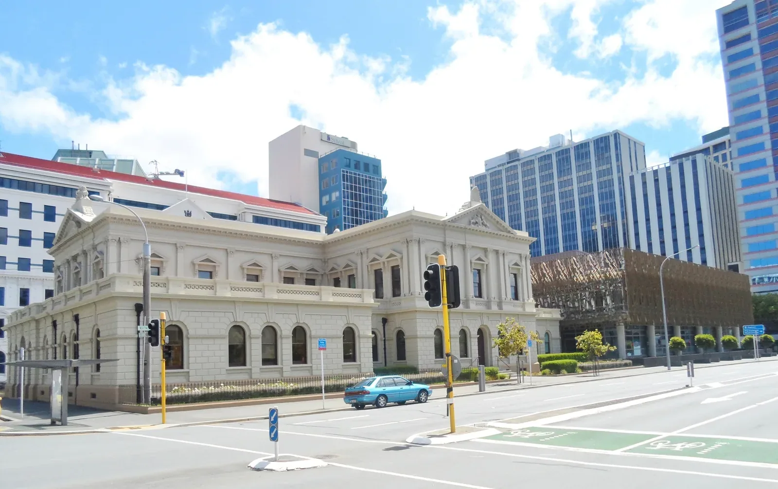 A white two-storey colonial-era court building on a Wellington street corner, with a modern building clad in a bronze lattice screen to its right and office towers behind