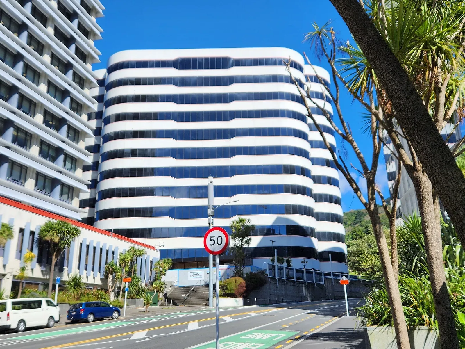 A tall curved white office tower with bands of blue windows above a sloping Wellington street, with a 50 km/h speed sign and palm trees in the foreground