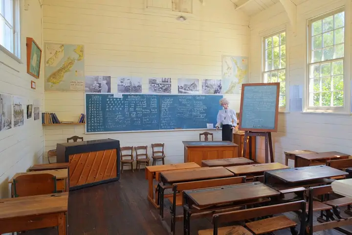 An old-style classroom with rows of wooden desks, blackboards covered in chalk writing, maps of New Zealand on the wood-panelled walls and a teacher figure standing at the front