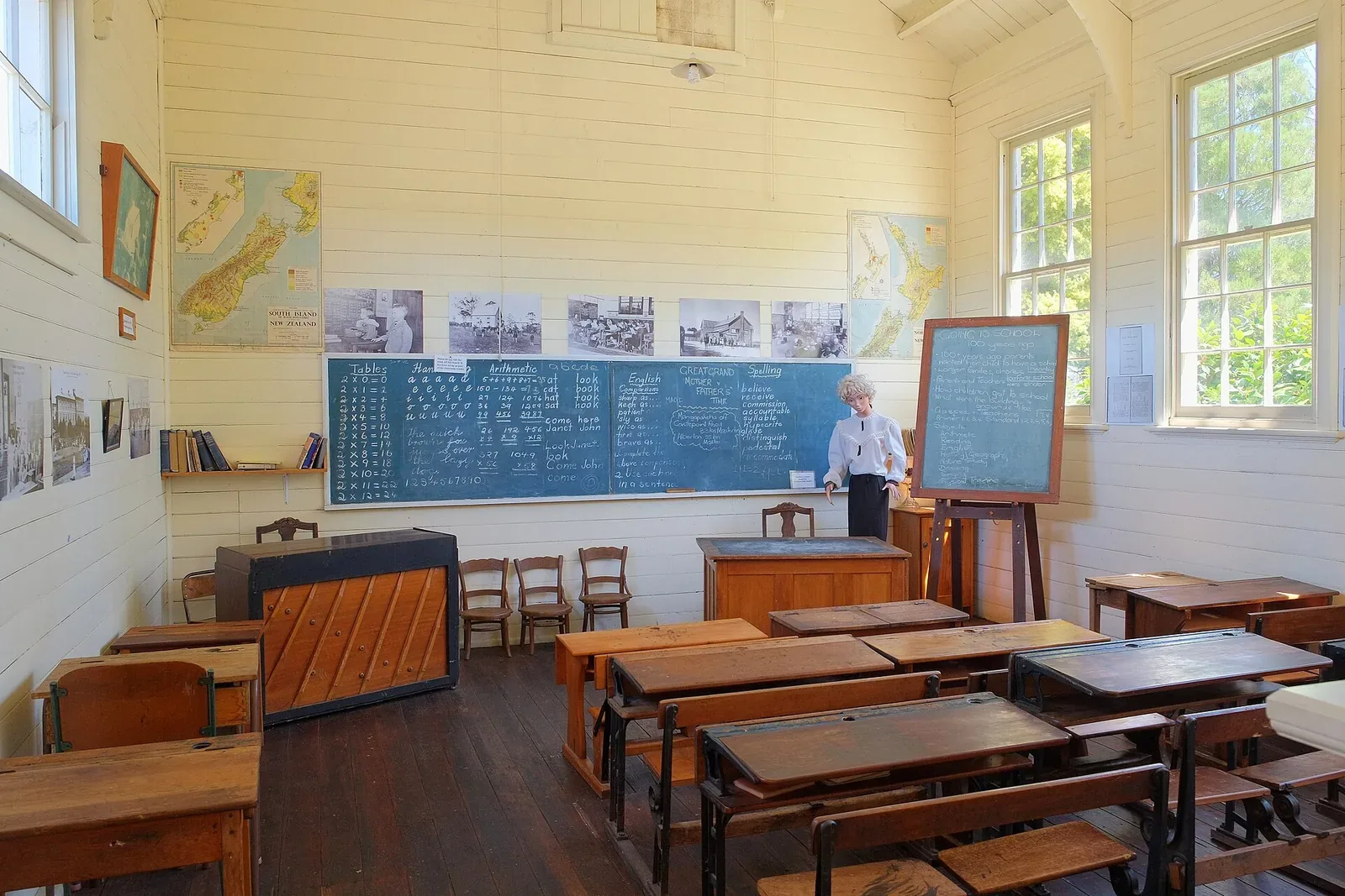 An old-style classroom with rows of wooden desks, blackboards covered in chalk writing, maps of New Zealand on the wood-panelled walls and a teacher figure standing at the front