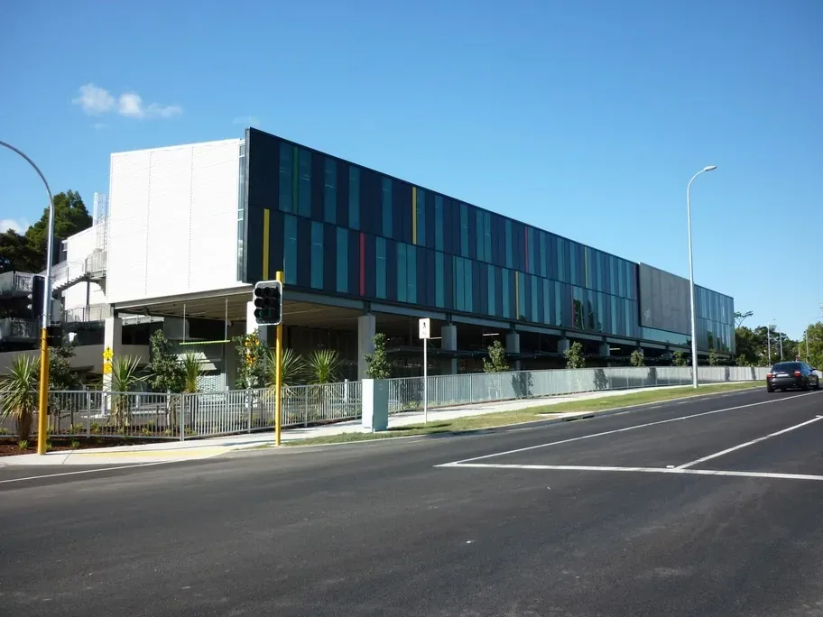 A modern three-storey school building with dark glass panels and coloured vertical stripes, beside a road under a clear blue sky