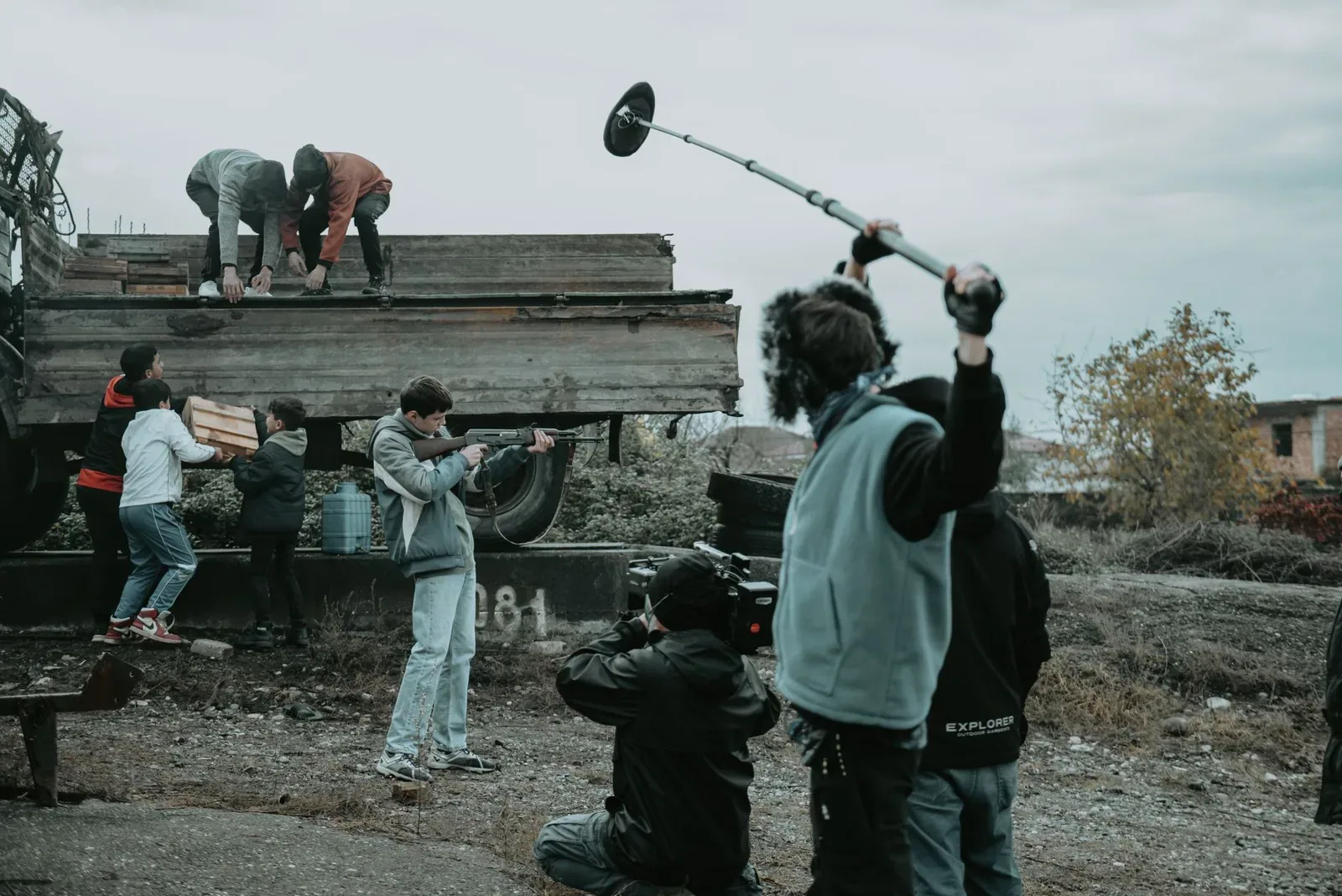 A film crew working outdoors on rough ground, with a boom operator holding a microphone overhead and a camera operator crouched filming people unloading a truck