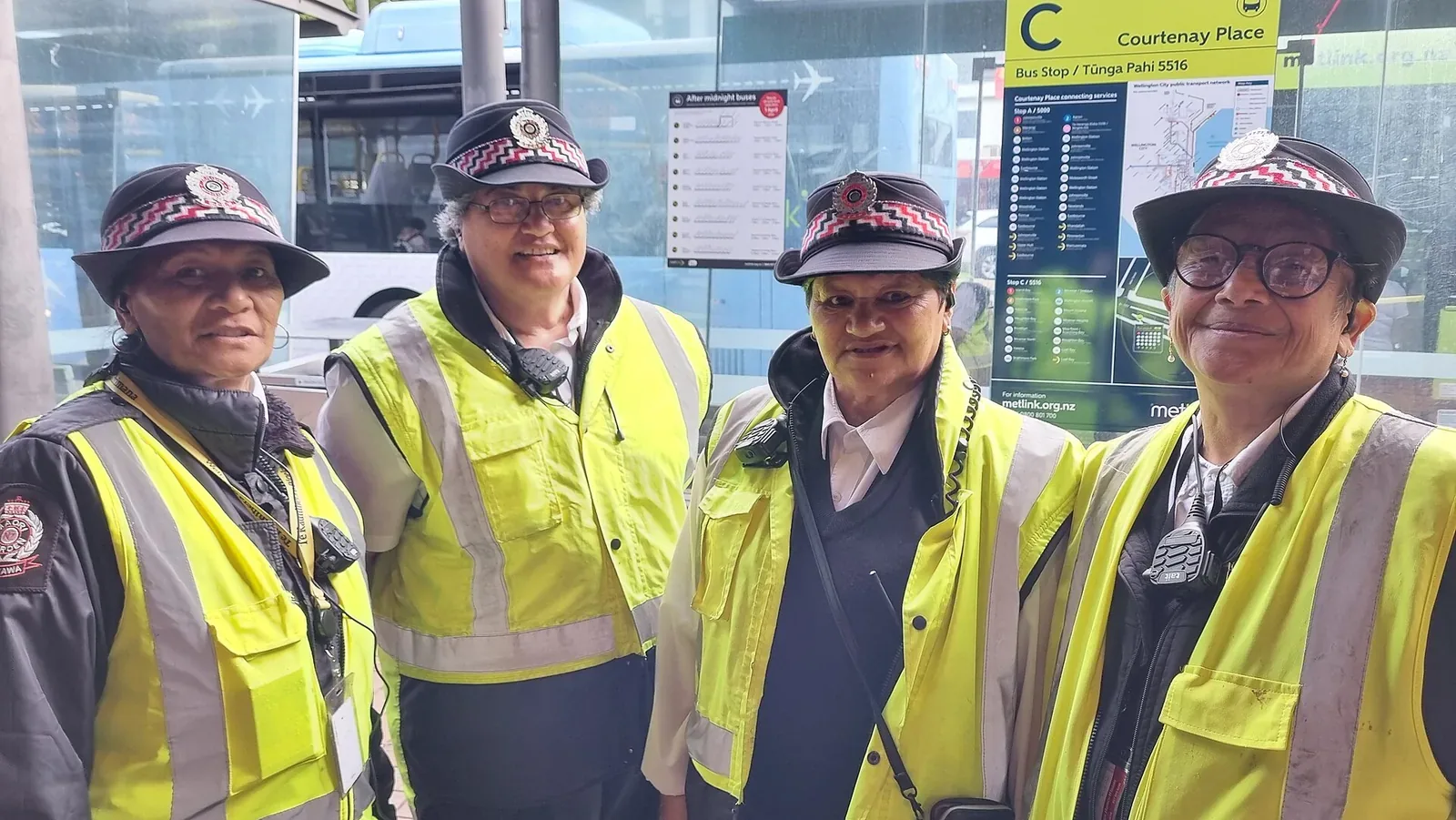 Four Māori Wardens in high-visibility yellow vests and peaked caps with chequered bands stand together at a Courtenay Place bus stop