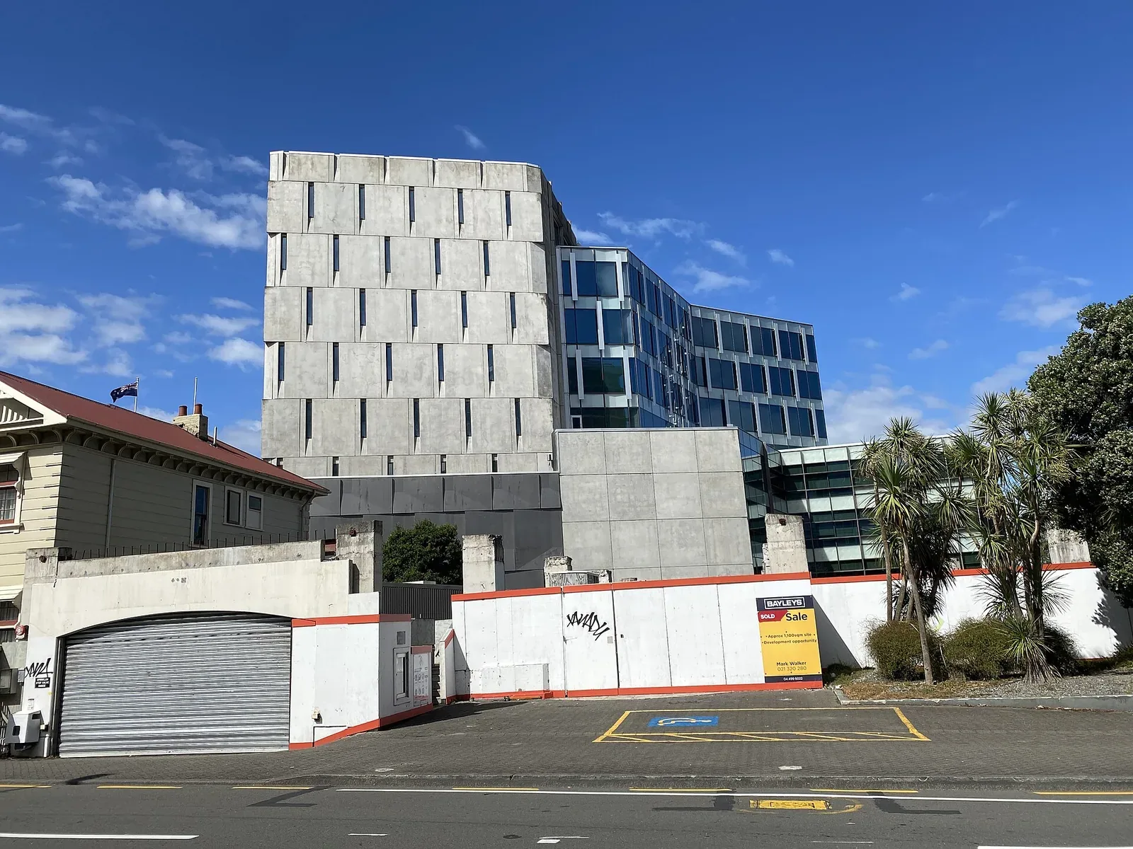 A grey concrete block building beside a blue glass office building, behind a white perimeter wall with a roller door, under a blue sky