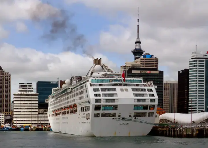 A large white cruise ship berthed at a wharf in front of city high-rises, with Auckland's Sky Tower rising behind it under a cloudy sky.