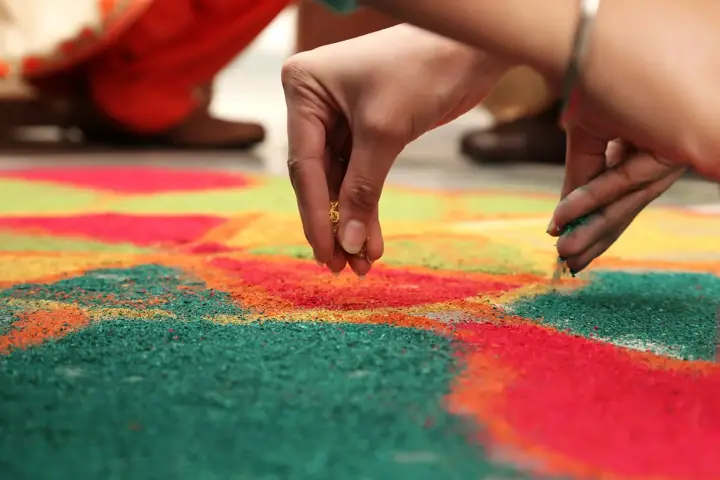 Close-up of hands sprinkling coloured powder to fill in a red, green, yellow and orange rangoli pattern on the floor