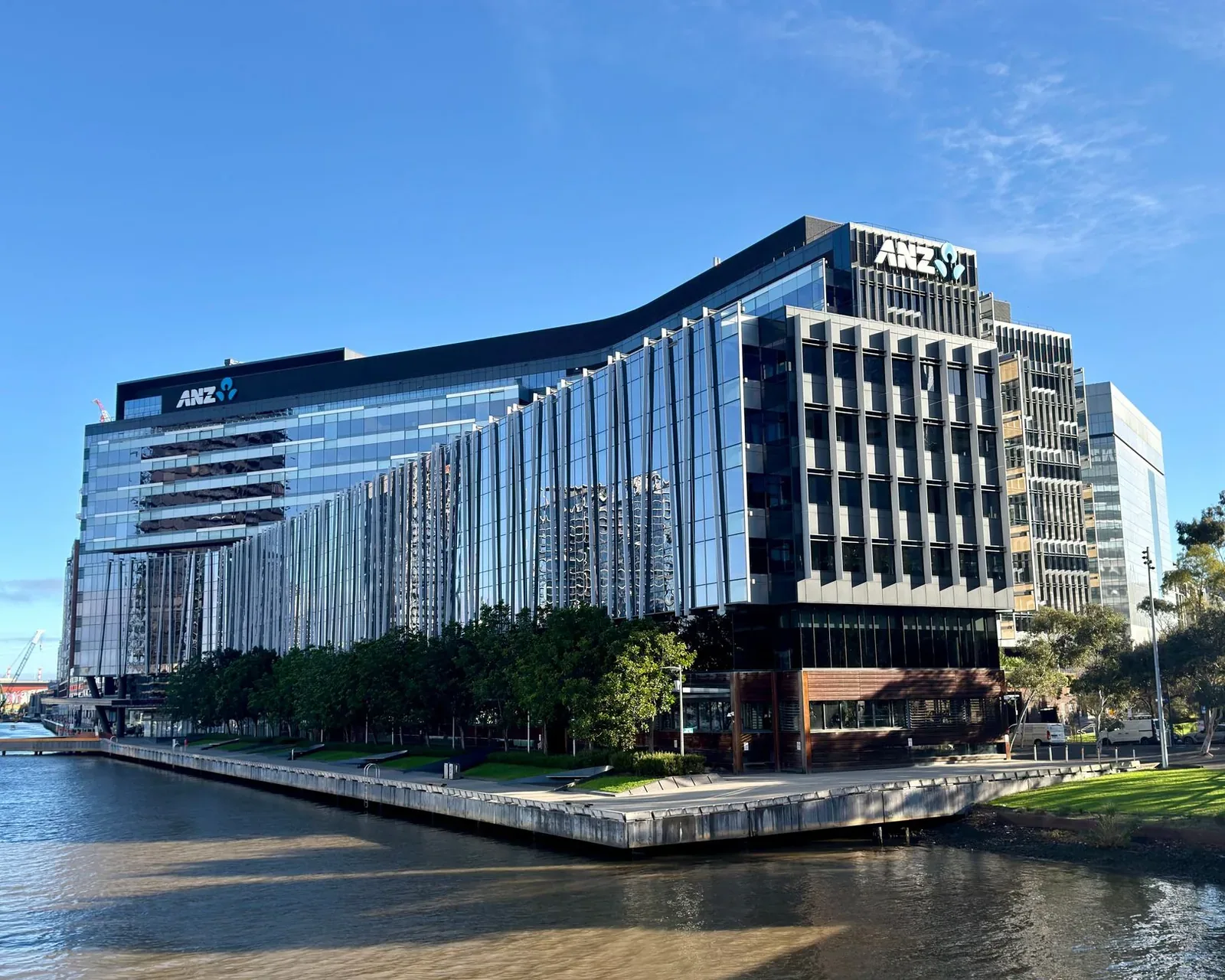 A large modern office building with a reflective glass facade and ANZ logos near the roofline, standing beside a waterway with a row of trees along the water's edge under a blue sky.
