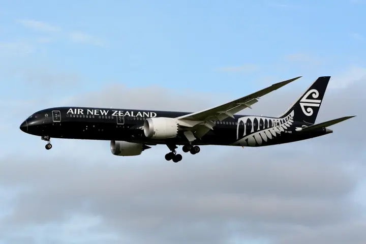 A black Air New Zealand Boeing 787-9 with a silver fern design on the rear fuselage flies with its landing gear down against a partly cloudy sky.