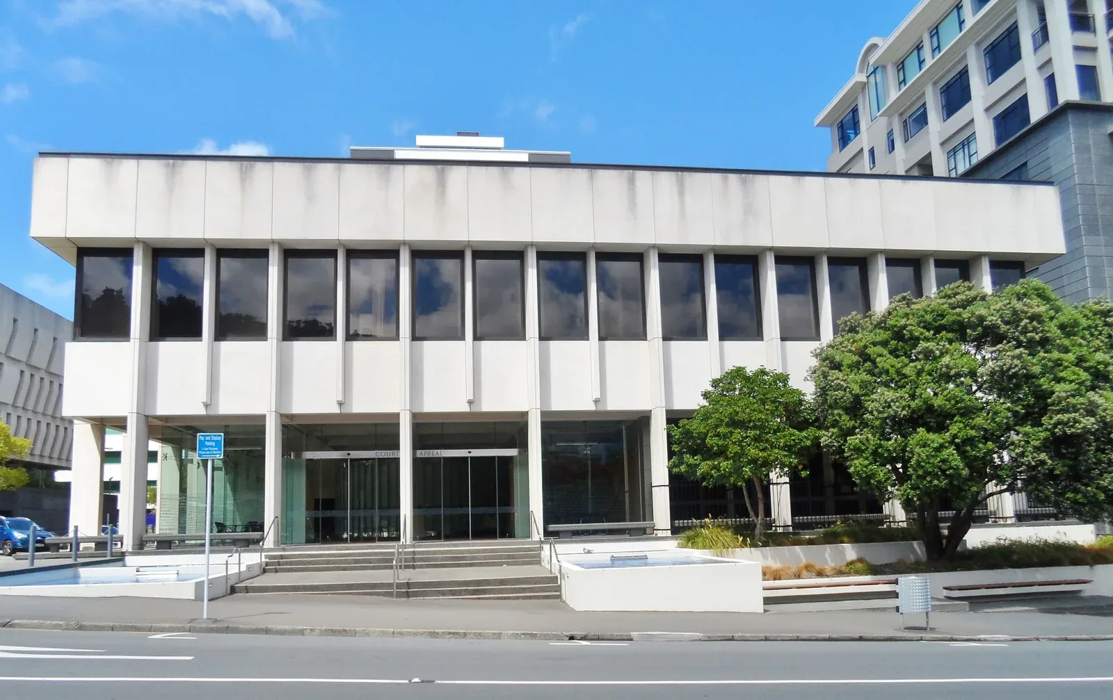 A pale, modernist four-storey building with tall vertical columns and dark windows, with steps and glass doors at street level and trees beside it under a blue sky