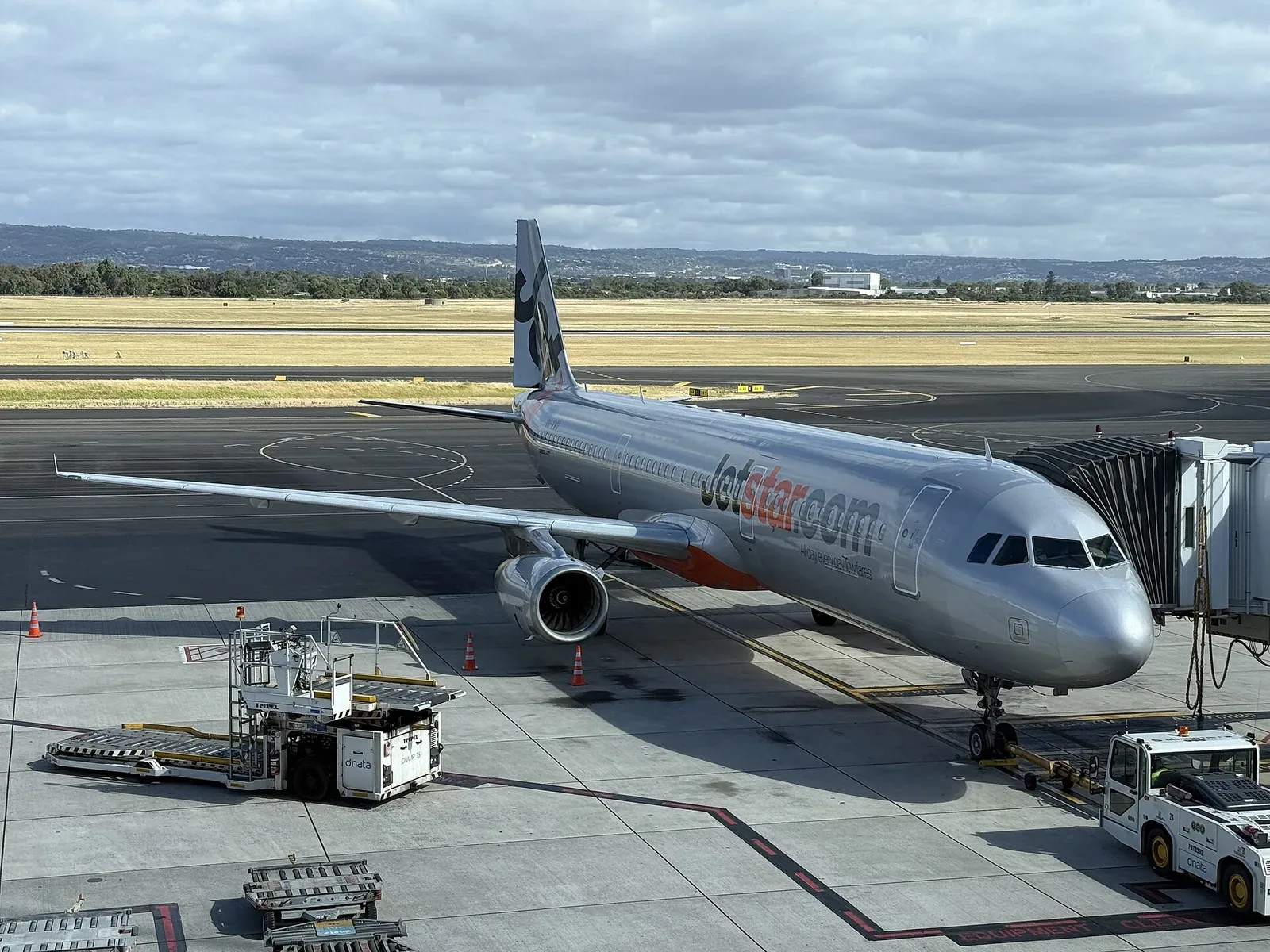 A silver Jetstar Airways Airbus A321 parked at an airport gate with an aerobridge attached and ground equipment beside it, under a cloudy sky