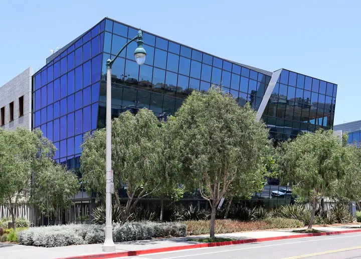 A modern office building with an angular blue glass front, behind a row of trees and a street lamp on a sunny day