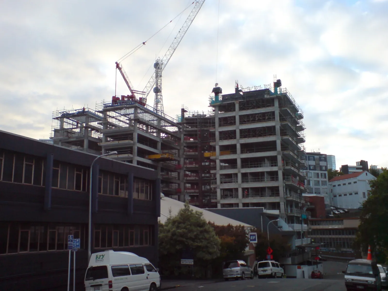 A multi-storey apartment building under construction, wrapped in scaffolding with tower cranes above, seen from a street with parked vans and cars