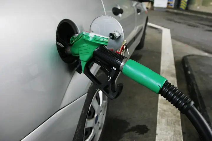 A green petrol pump nozzle inserted into the open fuel filler of a silver car at a petrol station
