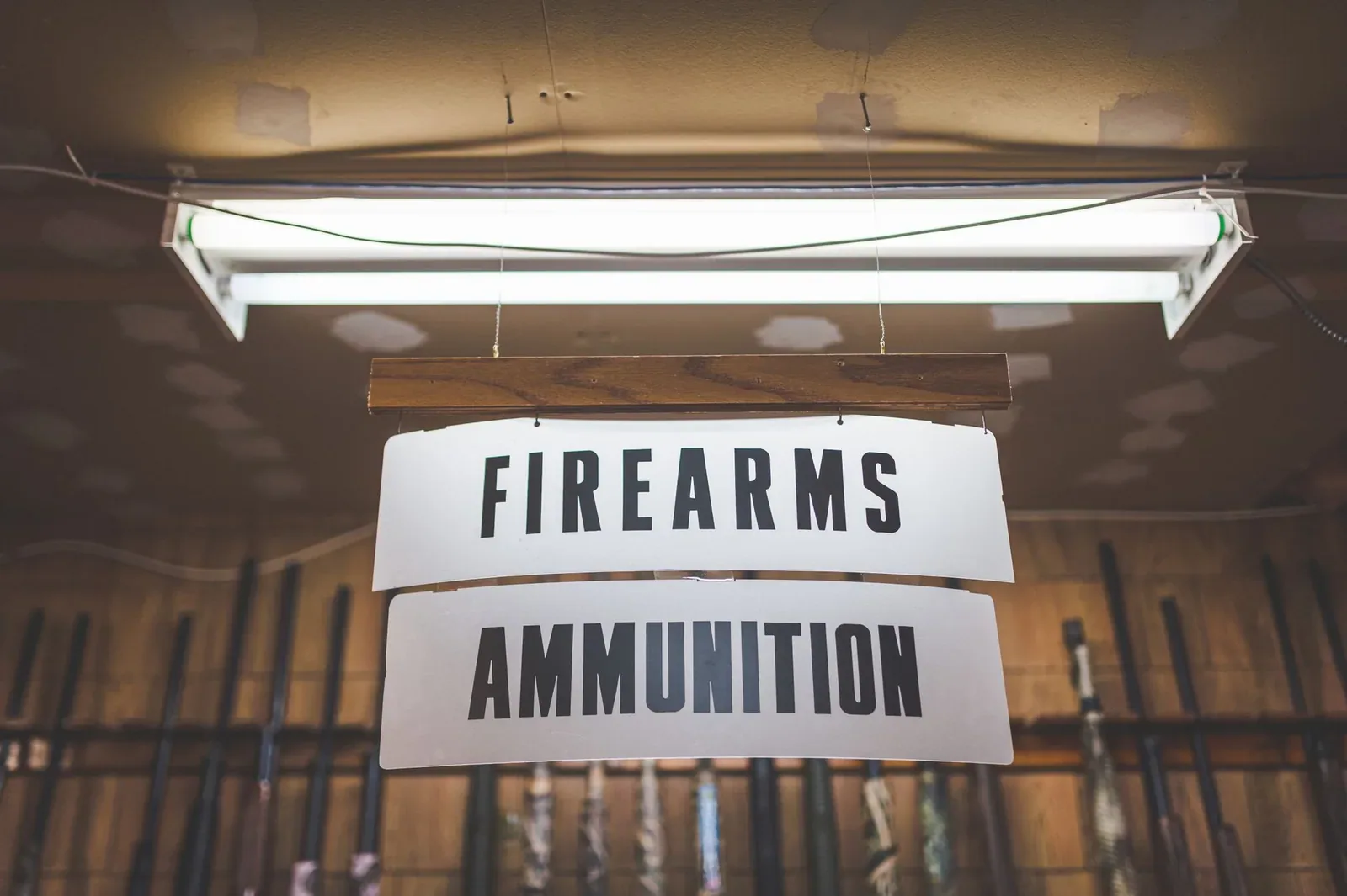 Two white hanging signs reading "Firearms" and "Ammunition" under a fluorescent light, with a rack of long guns behind them in a store