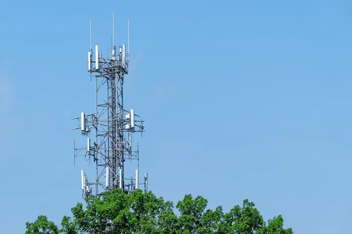 A lattice cellular telephone tower with several antennas rising above green treetops against a clear blue sky
