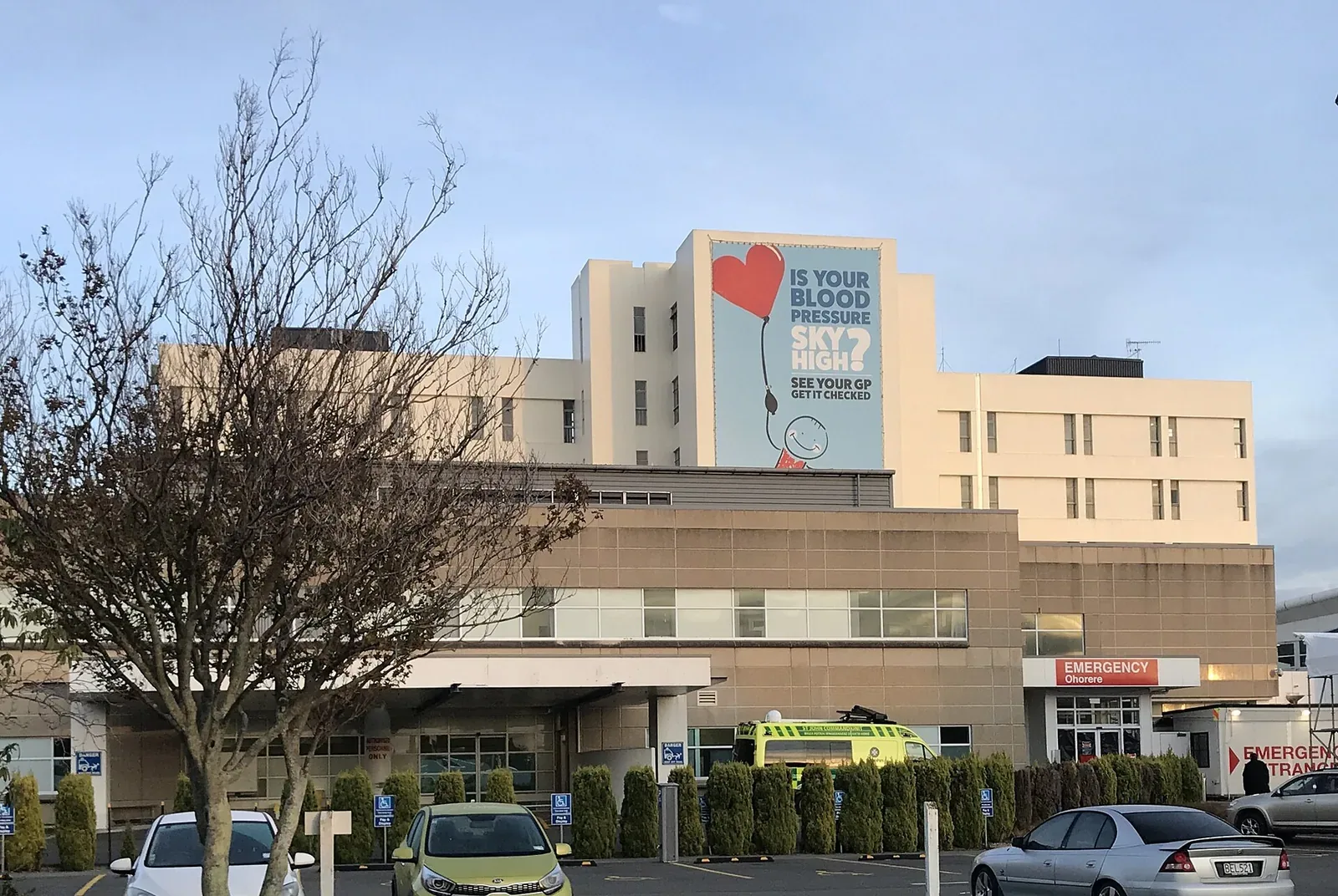 A multi-storey hospital building with a red 'Emergency' sign over the entrance on the right and an ambulance parked outside, cars in the car park in front