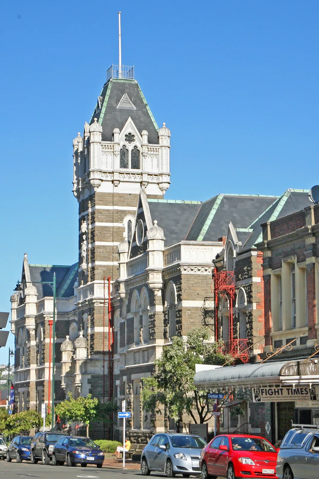 A tall Victorian stone courthouse with a central tower and slate roofs on a city street, with cars parked out front under a clear blue sky