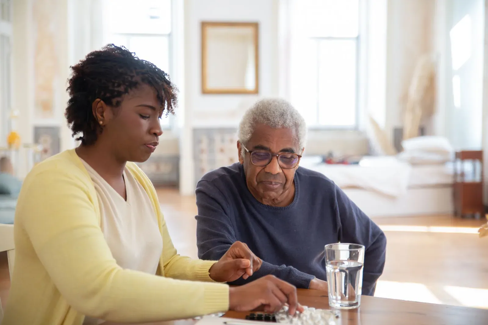A woman in a yellow cardigan sits at a table with an older man in glasses, helping him sort his medication, with a glass of water beside them