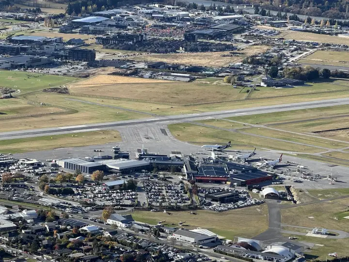 Aerial view of Queenstown Airport showing the terminal, parked aircraft, car parks and the runway, with surrounding town and fields