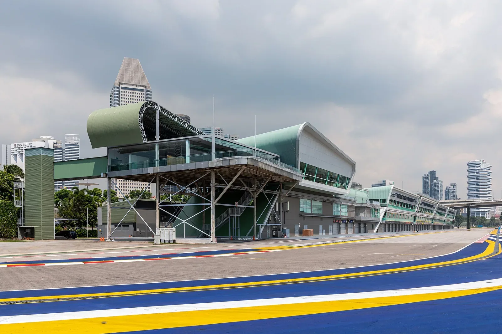 A long modern green-and-white pit building with a curved roof beside an empty racetrack with blue, white and yellow painted lines, city towers in the background under a cloudy sky