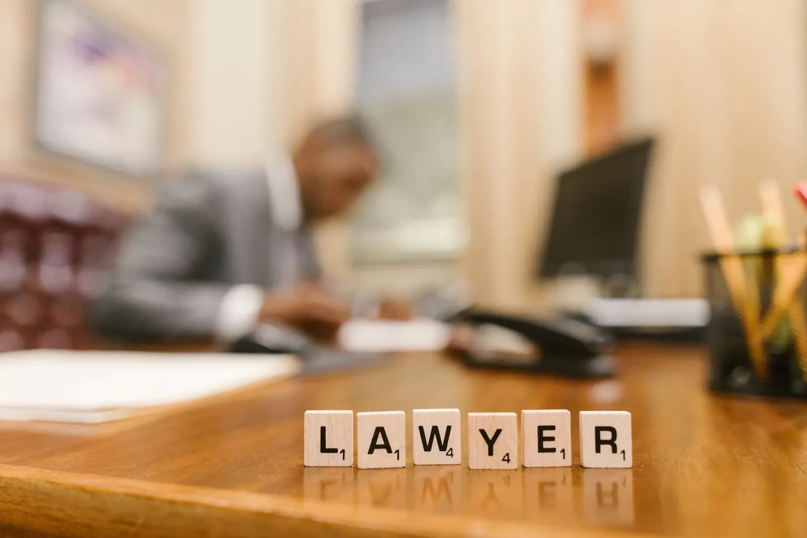 Wooden letter tiles spelling LAWYER on a wooden desk, with a blurred figure in a suit working at a desk in an office behind