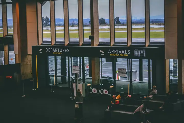An airport terminal hall with large windows looking onto the airfield, and Departures and Arrivals signs above glass doors. A few travellers sit on benches nearby.