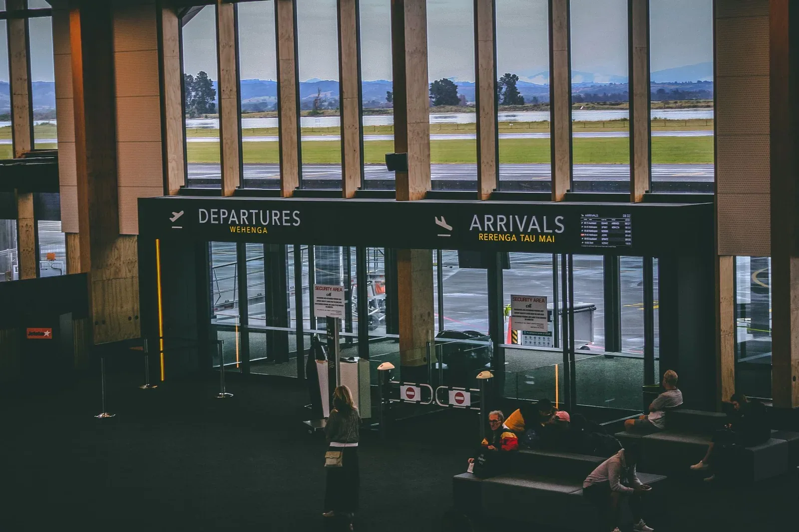 An airport terminal hall with large windows looking onto the airfield, and Departures and Arrivals signs above glass doors. A few travellers sit on benches nearby.